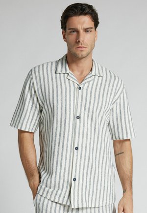 Mężczyzna ubrany w białą koszulę z krótkim rękawem i szarymi pionowymi paskami, stojący z jedną ręką w kieszeni na jednolitym tle.