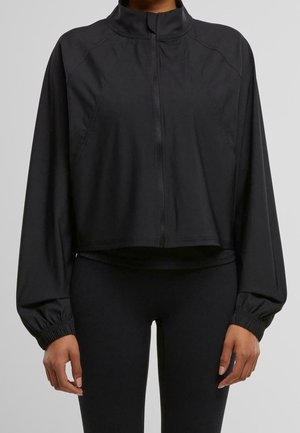 Žena v černé dlouhé zipové bundě s elastickými manžetami a černých legínách, zobrazená od krku k bokům.