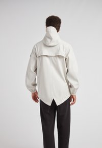 Lichtgrijze, waterdichte jas met een hoge kraag, capuchon en een verlengd achterpand. Lange mouwen hebben elastische manchetten; zijzakken zijn zichtbaar.