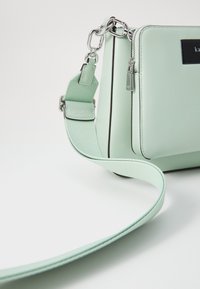 Borsa a tracolla sintetica verde chiaro con dettagli in argento, chiusura a zip e tracolla regolabile, caratterizzata da una texture liscia e un design minimalista.