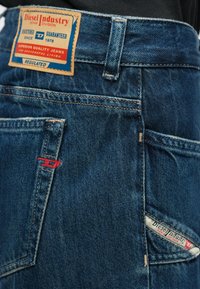 Sötétkék farmernadrág hátulnézetből, amely bőr márkajelzéssel, övlyukkal és piros-fehér Diesel Industry logókkal hímzett zsebekkel rendelkezik.