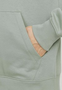 Haut à capuche vert avec une poche kangourou, des poignets côtelés et un tissu doux et texturé. Le design est simple et fonctionnel.