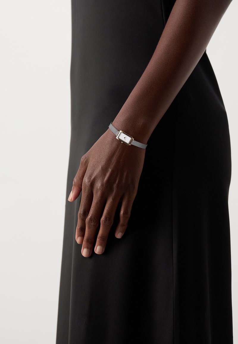 Rechter Arm und Hand einer Person, die ein schwarzes Kleid und eine rechteckige Armbanduhr mit grauem Armband trägt.