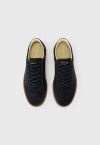 Adidași din suede de culoare bleumarin, cu șireturi de un albastru închis și talpă de cauciuc maro. Logo auriu pe limbă și texturi contrastante pe partea superioară pentru un plus de detaliu.