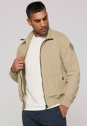 Mann richtet beige Reißverschlussjacke über weißem Hemd, trägt marineblaue Hose, vor schlichtem hellgrauem Hintergrund.
