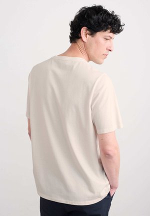 Licht beige T-shirt met korte mouwen, een ontspannen pasvorm, ronde halslijn en zachte textuur. De achterkant toont stikseldetails en een lichte draping.