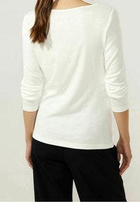 Frau, die ein weißes Langarmshirt und schwarze Hosen trägt, von hinten gezeigt, mit den Haaren zu einem lockeren Pferdeschwanz gebunden.