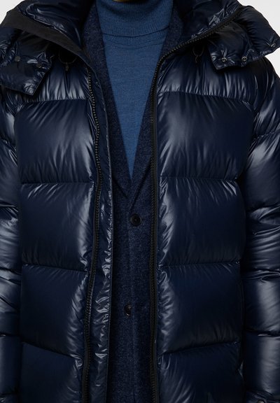 Navy blue puffer jacket with glossy finish, featuring a zip front, large quilted sections, and a hood. Worn over a dark blue sweater and vest.
