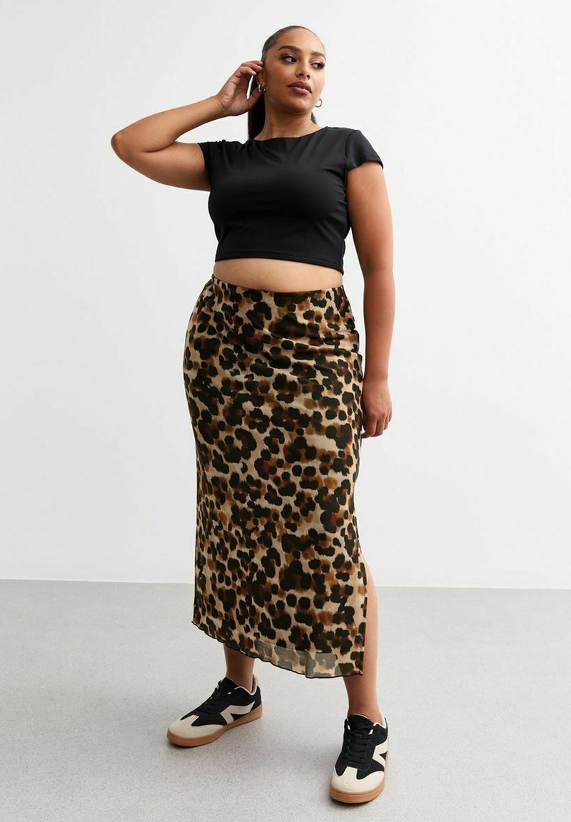 T-shirt noir court, jupe en satin à imprimé animal avec des taches vertes et brunes, fente sur le côté, associée à des baskets noires et beiges.