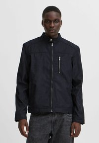 Chaqueta azul marino de tela suave con cuello alto, cremallera frontal y bolsillos laterales. Presenta un bolsillo en el pecho con cremallera y costuras mínimas.