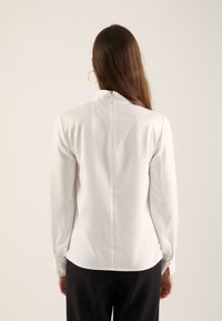 Femme aux longs cheveux bruns portant un chemisier blanc à manches longues avec fermeture éclair dans le dos et un pantalon noir, se tenant devant un fond uni.