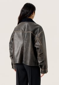 Brun læderjakke med en struktureret overflade, oversized pasform og kontrasterende sort corduroy-krave. Synlige syninger og sømme.