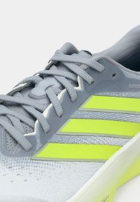 Jasnoszary but sportowy z trzema żółto-zielonymi paskami, chropowatą siateczką na cholewce, smukłym designem i szarymi sznurowadłami. Wykończony białą środkową podeszwą.