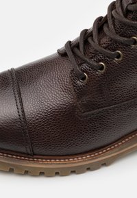 Brun texturerad läderstövel med mörkbruna snören, metallögon, sydd tådel och tjock gummisula med mönster.