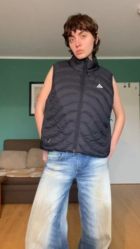 Czarna pikowana kamizelka z wysokim kołnierzem, zapinana na zamek, o prążkowanej teksturze; w zestawie z wyblakłymi, szerokimi jeansami w kolorze niebieskim.