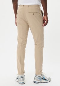 Beige Hosen mit taillierter Passform, ausgestattet mit zwei Gesäßtaschen und Knöpfen, abgerundetem Saum und einer glatten Stoffoberfläche. Kombiniert mit silbernen Sportschuhen.