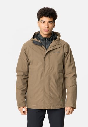 Junger Mann mit lockigem Haar, der eine beige wasserdichte Jacke über einer schwarzen Zip-Up-Schicht trägt, steht vor einem schlichten hellen Hintergrund.