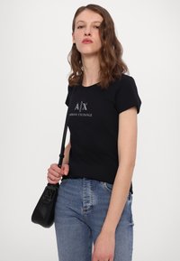 Tricou negru cu mânecă scurtă, cu logo-ul "A/X ARMANI EXCHANGE" imprimat în argintiu, asortat cu blugi albaștri și o geantă mică de umăr neagră.