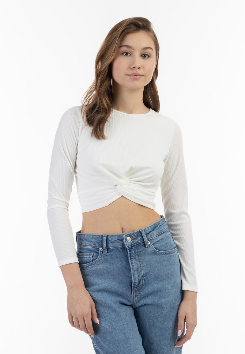 Crop top bianco a maniche lunghe con texture a coste e dettaglio con nodo anteriore, abbinato a jeans azzurri a vita alta.