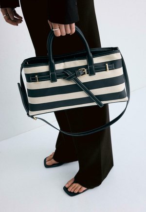 Hånd, der holder sort/hvid stribet rektangulær håndtaske med guldlås, iført sorte bukser og sorte sandaler med åbne tæer på hvidt gulv.