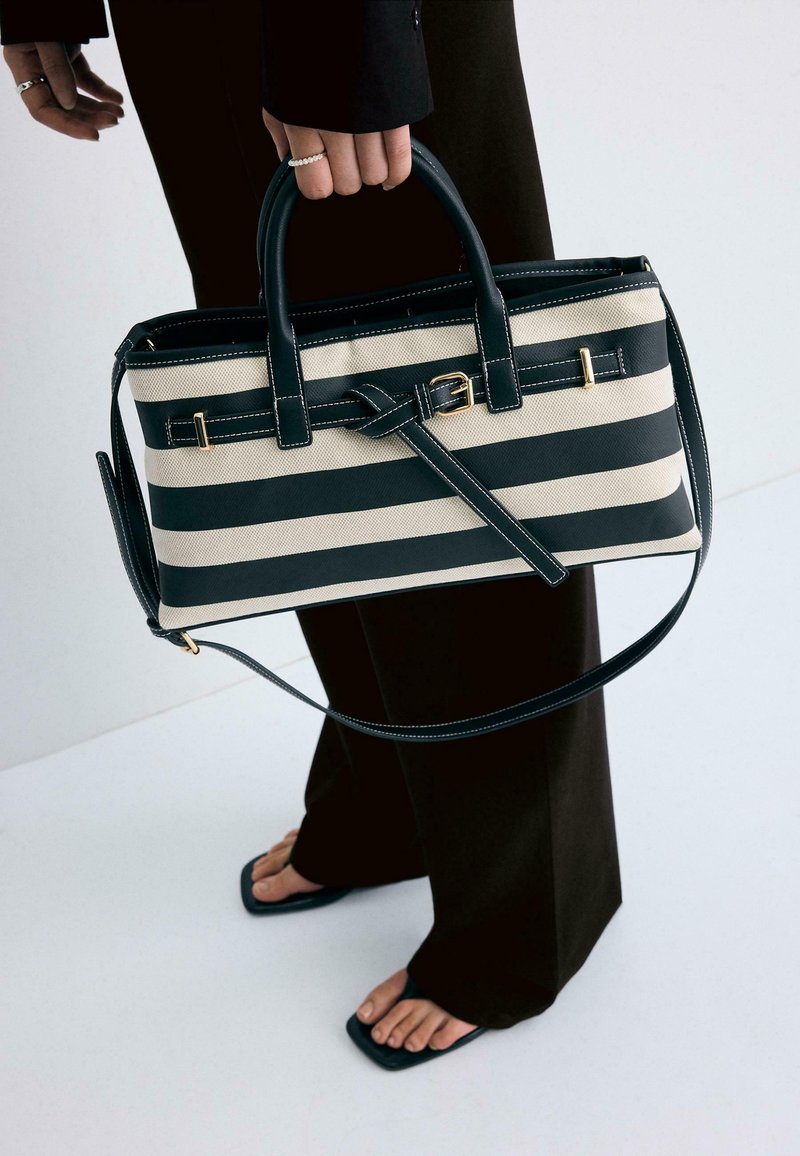 Main tenant un sac à main rectangulaire rayé noir et blanc avec une boucle dorée, portant un pantalon noir et des sandales noires à bout ouvert sur un sol blanc.