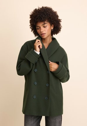 Femme aux cheveux bouclés portant un manteau croisé vert foncé, ajustant le col, debout devant un fond beige uni.