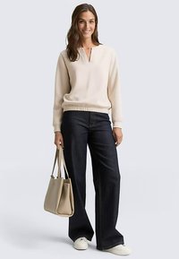 Maglione beige chiaro con scollo a V, polsini e fascia in vita a costine, abbinato a jeans ampi blu scuro e borsa beige. Scarpe da ginnastica bianche.