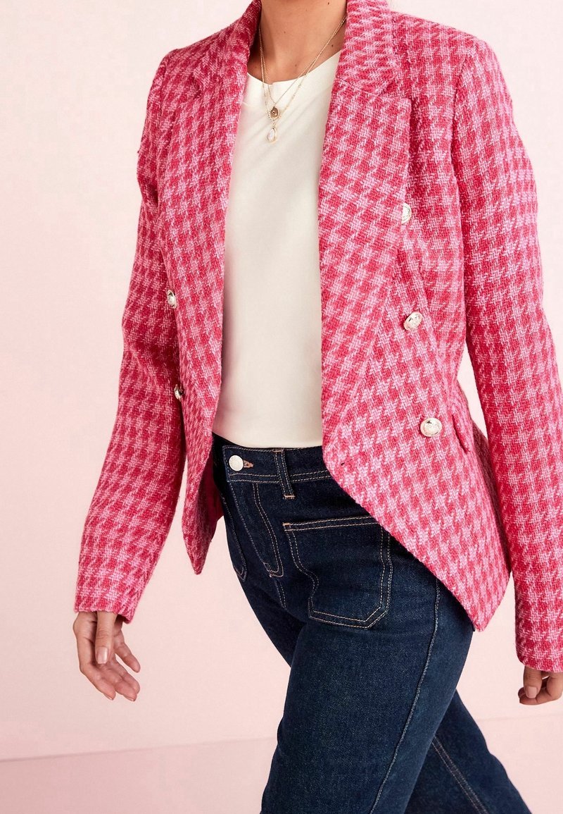 Femme portant un blazer croisé rose à motif pied-de-poule, un top blanc, un jean bleu foncé et des colliers dorés superposés, marchant sur un fond clair.