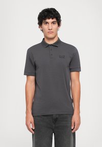 Young man with short curly hair wears dark gray short-sleeve polo shirt and black jeans, standing against a plain white background.