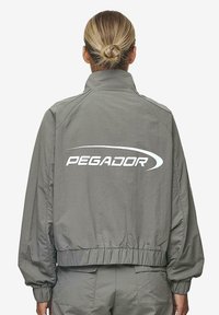 Hallide bomber-jakk, millel on kõrge krae, elastne vöökoht ja mansetid, tagaküljel on suur valge "PEGADOR" logo. Sujuv tekstuur.