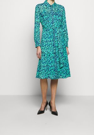 Kvinde iført en langærmet midikjole med blåt og mørkeblåt blomsterprint, krave, bælte og sorte spidse høje hæle, stående på en grå gulv.