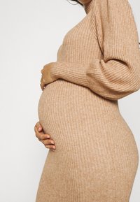 Robe en tricot beige clair avec texture côtelée, silhouette ajustée et manches longues. Les mains du modèle reposent sur un ventre visiblement enceinte.