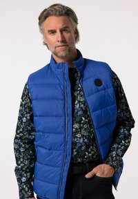 Blå isolerad väst med hög krage, dragkedjestängning och en rund svart logotyp. Buren över en mörkblommig mönstrad skjorta.