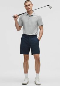 Heren golfkleding met een lichtgrijze polo met korte mouwen en marineblauwe shorts. Gecombineerd met een golfclub en witte golfschoenen.