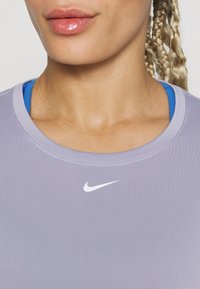 Světlé fialové sportovní tričko s kulatým výstřihem. Obsahuje bílé logo Nike na hrudi, navrstvené přes modré tílko. Hladká textury.