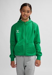 Grøn zip-hoodie med hætte, lavet af blødt stof. Har kengurulommer og "Hummel" logo broderet på brystet.
