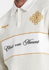 Grijze poloshirt met een witte horizontale band met zwarte tekst "Elevate EVERY Moment" en een gouden geborduurde bloemenontwerp. Witte kraag.