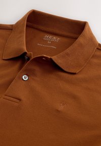 Rudos spalvos polo marškinėliai įprasto silueto, pagaminti iš tekstūruotos medžiagos, su apykakle ir vienu sidabriniu mygtuku. Ant krūtinės subtiliai išsiuvinėtas logotipas.