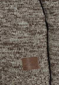 Knitted sweater featuring a brown and cream speckled pattern. Includes a brown leather tag with text and decorative elements.