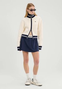 Cremefarbener Strick-Jacke mit Reißverschluss und marineblauen Akzenten, kombiniert mit einem marineblauen Faltenrock. Das Modell trägt schwarze Sonnenbrillen und weiße Sportschuhe.