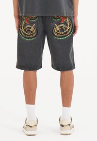 Tumman farkun shortsit, joissa on kirkkaan värisiä käärmeitä brodeerattuna takataskuissa. Suora leikkaus ja polven pituinen malli.