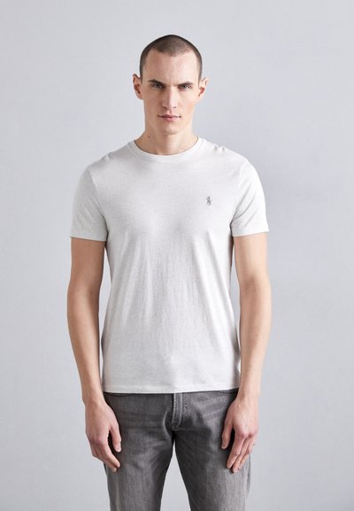 T-shirt en coton gris clair à manches courtes, col rond et petit logo brodé sur la poitrine gauche. Porté avec un jean gris.