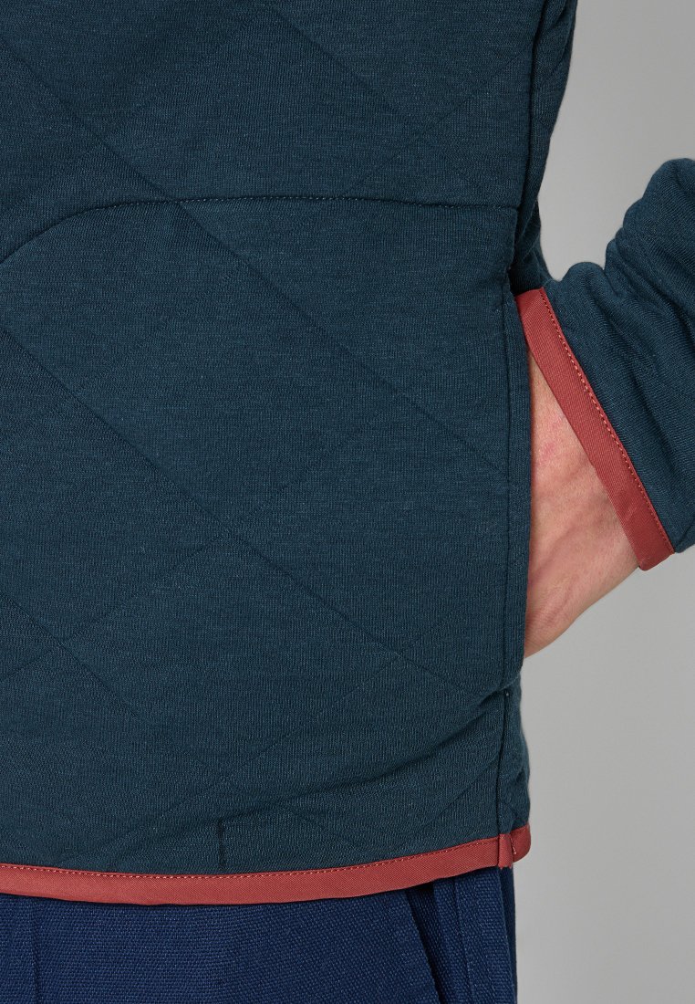 Tessuto trapuntato verde scuro con una vestibilità regolare, caratterizzato da un bordo rosso a contrasto lungo i polsini e l'orlo, che presenta un design elegante.