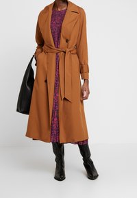 Abrigo trench marrón con cintura ceñida, mangas largas y detalles de botones, usado sobre un vestido floral. Bolso de hombro negro y botas hasta la rodilla.