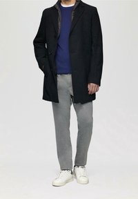 Man in een zwarte jas over een blauwe trui, grijze broek en witte sneakers, staand met één hand in zijn zak tegen een effen achtergrond.