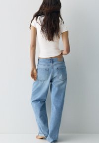 Hosszú barna hajú nő, fehér testhezálló pólót és bő, világoskék farmert visel, mezítláb áll egy sima világosszürke fal felé fordulva.