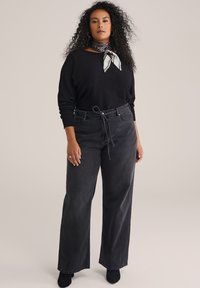 Fekete, hosszú ujjú felső, magasan szabott, bő szárú fekete farmerral párosítva. Az öltözetet egy kontrasztos mintázatú sál díszíti, amely a nyakon van megkötve.