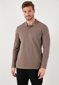 Camisa polo de manga larga, marrón, con tejido texturizado. Presenta un cuello clásico y una tapeta de tres botones, combinada con pantalones negros.