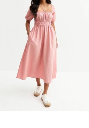Vrouw draagt een roze midi-jurk met pofmouwen en een geplooide bovenkant, gecombineerd met witte en beige sneakers, staand tegen een effen achtergrond.