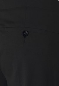 Zwarte broek met een gladde textuur, met een enkele achterzak die beveiligd is met een ronde zwarte knop. Subtiele stiksels zijn zichtbaar.